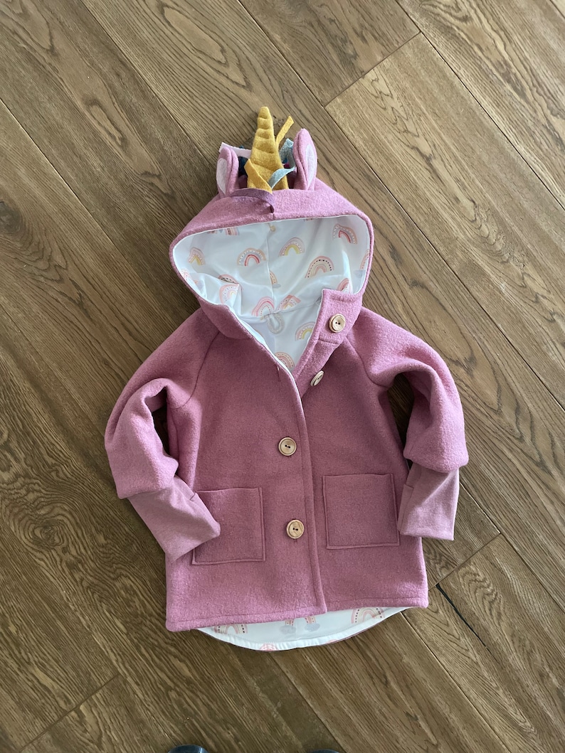 May include: A pink hooded jacket with a unicorn horn on the hood. The jacket has two pockets and a button closure. The lining of the hood is white with a rainbow pattern.