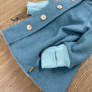 Puede incluir: Una chaqueta con capucha azul turquesa con botones de madera y forro de polar azul claro. La chaqueta tiene un solo bolsillo en el lado izquierdo.