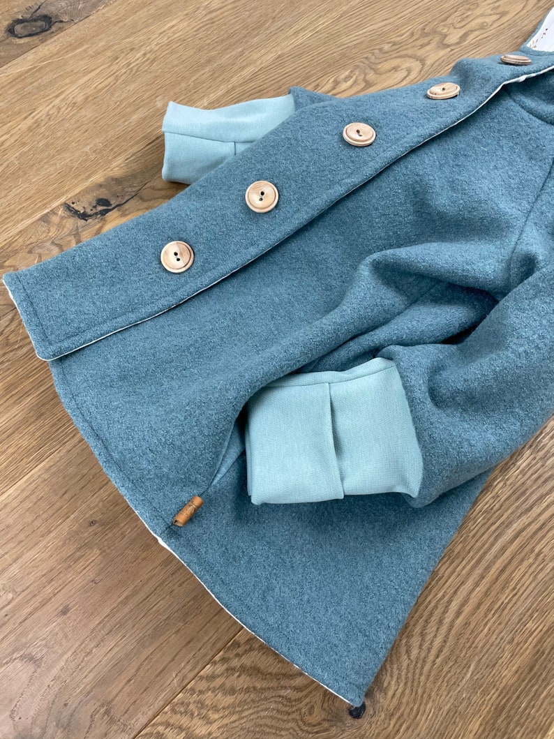 Puede incluir: Una chaqueta con capucha azul turquesa con botones de madera y forro de polar azul claro. La chaqueta tiene un solo bolsillo en el lado izquierdo.