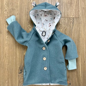 Puede incluir: Una chaqueta azul turquesa con capucha y forro estampado en blanco y marr&oacute;n. La chaqueta tiene cierre de botones y dos bolsillos. La capucha tiene dos peque&ntilde;as orejas en la parte superior.