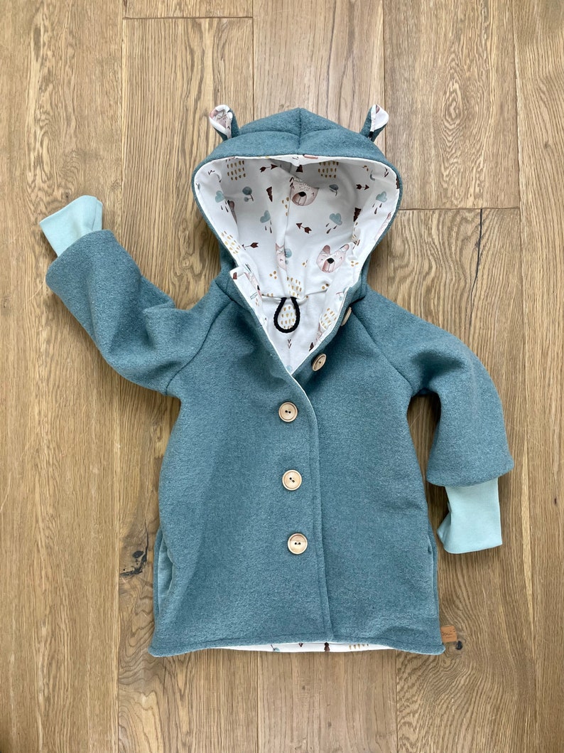 Puede incluir: Una chaqueta azul turquesa con capucha y forro estampado en blanco y marr&oacute;n. La chaqueta tiene cierre de botones y dos bolsillos. La capucha tiene dos peque&ntilde;as orejas en la parte superior.