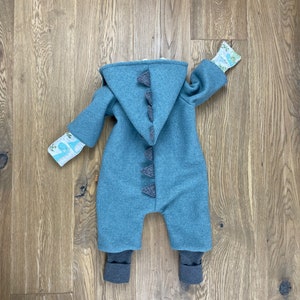 Puede incluir: Un mono azul con capucha y un diseño de cola de dinosaurio. El mono tiene puños marrones y un tejido estampado blanco y verde en las mangas.