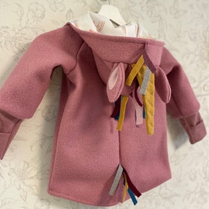 May include: A pink hooded jacket with a unicorn tail made of colorful fabric strips. The hood has a white lining with a rainbow pattern.