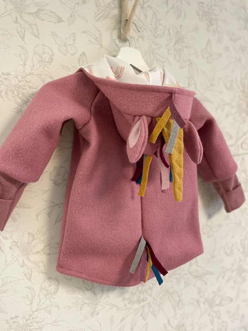 May include: A pink hooded jacket with a unicorn tail made of colorful fabric strips. The hood has a white lining with a rainbow pattern.