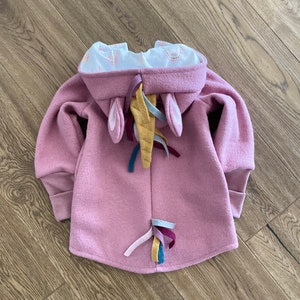 May include: A pink hooded sweatshirt with unicorn ears and a rainbow tail. The hood is lined with a white fabric with a small pink and white pattern.