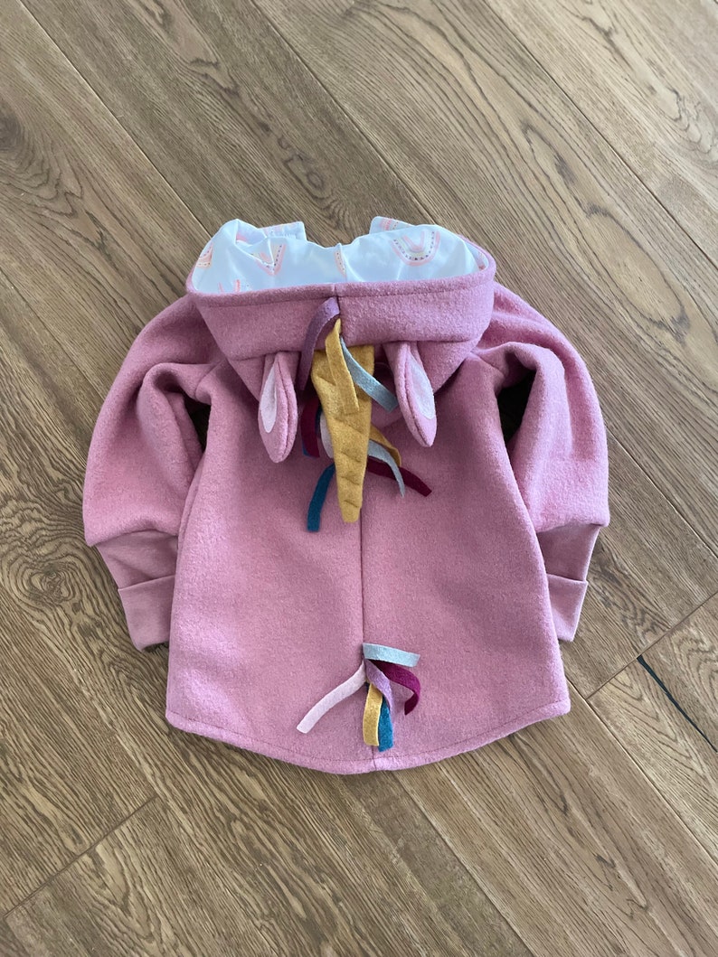 May include: A pink hooded sweatshirt with unicorn ears and a rainbow tail. The hood is lined with a white fabric with a small pink and white pattern.