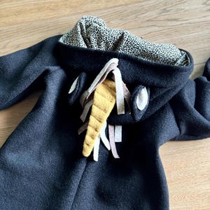 Puede incluir: Mono negro con capucha y una aplicaci&oacute;n de fieltro amarillo y marr&oacute;n en forma de cara de animal. La capucha est&aacute; forrada con un tejido de estampado de leopardo negro y blanco.