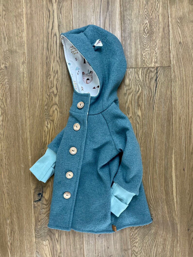 Puede incluir: Una chaqueta azul turquesa con capucha y forro estampado en blanco y marr&oacute;n. La chaqueta tiene cierre de botones y mangas largas con pu&ntilde;os azul turquesa.