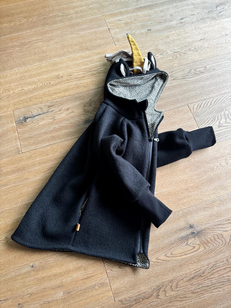 Puede incluir: Una chaqueta negra con capucha con un dise&ntilde;o de unicornio en la capucha. La capucha est&aacute; forrada con un estampado de animal negro y blanco.