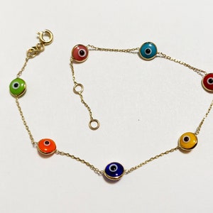 Może przedstawiać: Złota bransoletka z łańcuszkiem z siedmioma kolorowymi wisiorkami w kształcie oka. Wisiorki są okrągłe i mają białą źrenicę w środku. Bransoletka ma delikatny design i byłaby świetnym prezentem.