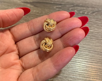 May include: A pair of gold knot-shaped earrings. The earrings are small and have a textured surface.