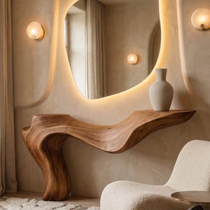 May include: A wall-mounted console table crafted from natural wood with a fluid, organic, wave-like shape. Above the table hangs an asymmetrical, backlit mirror. A matte beige vase sits on the table surface, and a textured cream chair is nearby.