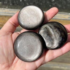 May include: Three polished, round, silver sheen obsidian stones are held in a hand. The stones have a dark, reflective surface with a metallic shimmer. The stones vary slightly in their patterns and coloration.