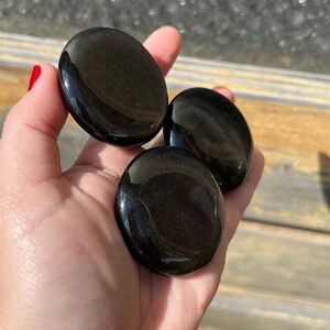 May include: Three polished, black obsidian palm stones are held in a hand. The stones are smooth, oval-shaped, and reflect light, showcasing a glossy finish. The stones are a deep, solid black color.