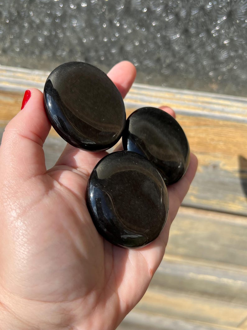 May include: Three polished, black obsidian palm stones are held in a hand. The stones are smooth, oval-shaped, and reflect light, showcasing a glossy finish. The stones are a deep, solid black color.