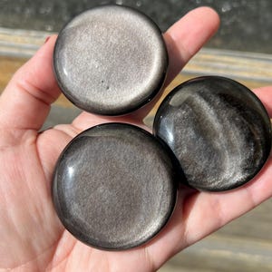 May include: Three polished, round, obsidian stones with a metallic sheen are held in a hand. The stones have a dark, reflective surface with swirling patterns and a silver-gray shimmer. The stones are approximately 2 inches in diameter.