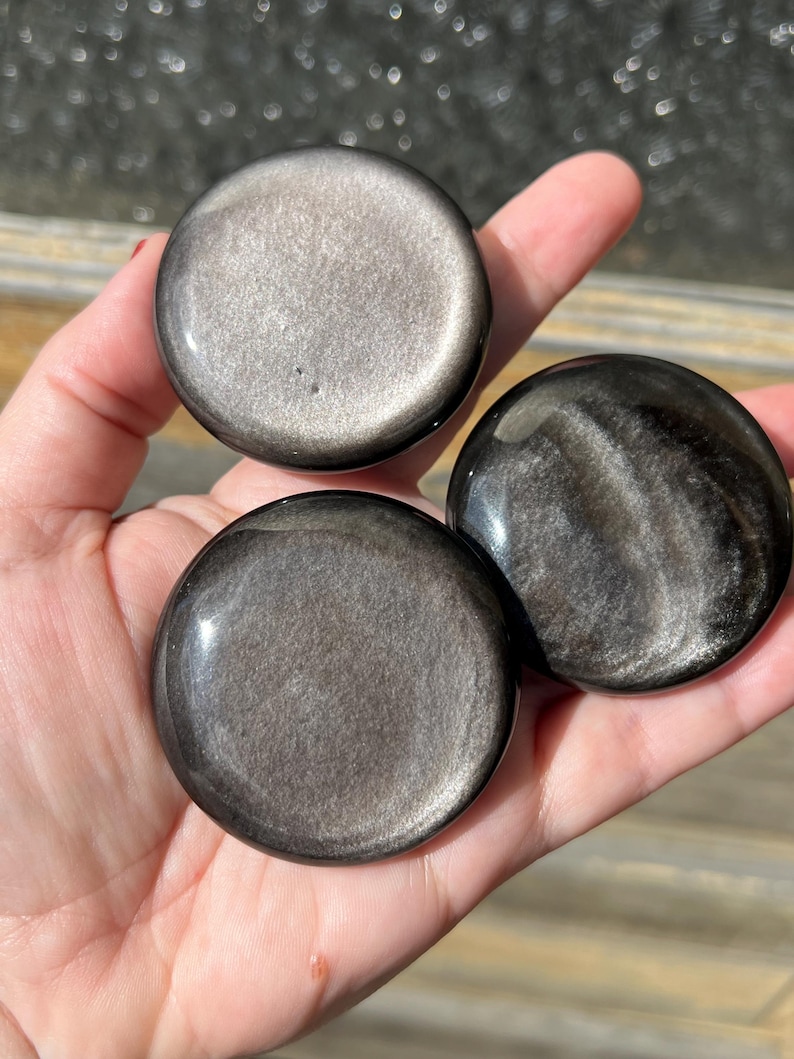 May include: Three polished, round, obsidian stones with a metallic sheen are held in a hand. The stones have a dark, reflective surface with swirling patterns and a silver-gray shimmer. The stones are approximately 2 inches in diameter.
