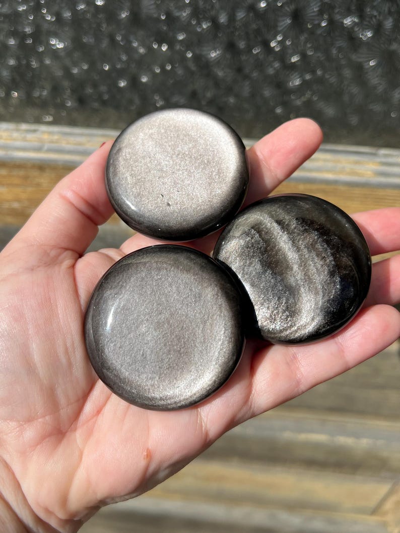 May include: Three polished, round, silver sheen obsidian stones are held in a hand. The stones have a dark, reflective surface with a metallic shimmer. The stones vary slightly in their patterns and coloration.