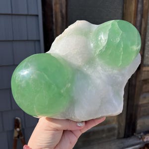 May include: A large, raw, natural fluorite crystal. The crystal features two prominent, rounded, light green spheres atop a white, opaque base. The stone is held in a hand, showcasing its size and texture. The background is a blurred outdoor scene.