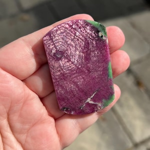 May include: A rectangular, polished gemstone with a deep magenta hue, displaying a unique, textured pattern. The stone has areas of green and white, adding contrast. The gemstone is held in a hand, showcasing its size and shape.