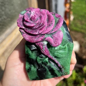 May include: A hand-carved stone sculpture featuring a prominent magenta rose blossom with layered petals resting atop a base of deep green zoisite with black hornblende inclusions, showcasing a carved stem and leaves extending downward.