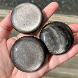 May include: Three polished, round obsidian stones with a metallic sheen are displayed. The stones vary in their silver and black coloration, with swirling patterns visible on some. The smooth, reflective surfaces catch the light, creating a captivating display.