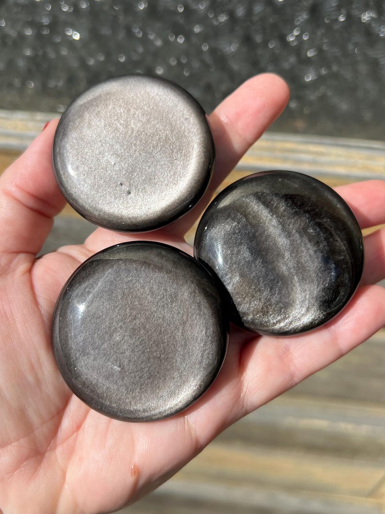 May include: Three polished, round obsidian stones with a metallic sheen are displayed. The stones vary in their silver and black coloration, with swirling patterns visible on some. The smooth, reflective surfaces catch the light, creating a captivating display.