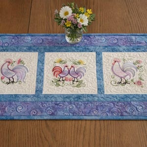 May include: A rectangular, quilted table runner with a blue and white color scheme. The center features three embroidered panels with rooster designs in shades of purple, red, and green. The border has a swirling pattern.