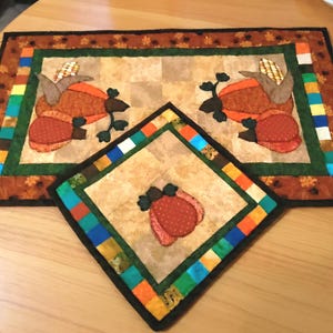 May include: A quilted placemat and matching coaster set featuring a fall harvest theme. The placemat has a brown border with a colorful striped accent and a central design of two turkeys with corn. The coaster has a similar design with a single pumpkin.
