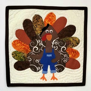 May include: A square fabric wall hanging featuring a brown turkey appliqué wearing blue denim-style overalls. The turkey has googly eyes, a red wattle, and an orange beak. The tail feathers are made of various patterned fabrics in brown, orange, and green.