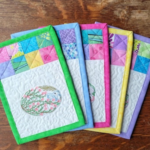 May include: Five colorful quilted pot holders with embroidered designs. Each pot holder features a different patchwork pattern with a white background and a green, blue, pink, yellow, or purple border.