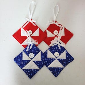 May include: Four quilted ornaments in red and blue, with white accents and button details. Two are red with white triangles, and two are blue with white star patterns. Each has a white ribbon for hanging.
