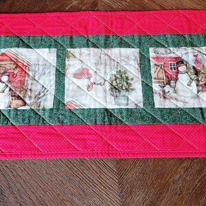 May include: A red quilted table runner with a green border and three winter-themed fabric squares featuring snowmen, houses, and trees.