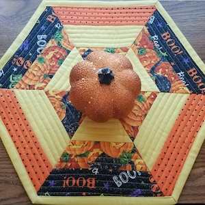 May include: A hexagonal table runner with a yellow, orange, and black patchwork design. The center features a glittery orange pumpkin with a black stem. The fabric has a "BOO!" pattern and black stars.