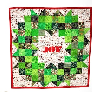 May include: A red, white, and green patchwork quilt with a starburst design. The center of the quilt features the word "JOY" in red.