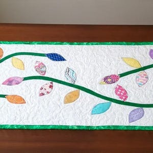 May include: A rectangular quilted table runner with a white background featuring a swirling green vine pattern. The design includes various colorful fabric leaves in shades of orange, yellow, pink, blue, and purple, finished with a green border.