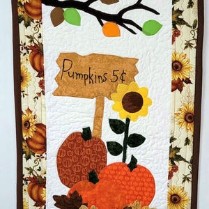 May include: A quilted wall hanging with a fall theme. It features a central panel with pumpkins, a sunflower, and a sign that reads "Pumpkins 5¢". The border has sunflowers and pumpkins. The color palette includes orange, brown, green, and yellow.