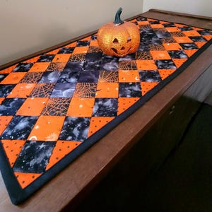 May include: A Halloween-themed table runner with a patchwork design of orange and black squares. The runner features spiderwebs, stars, and polka dots. A glittery orange pumpkin with a carved face sits on top of the runner.