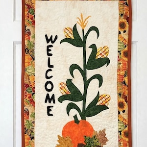 May include: A rectangular fabric wall hanging featuring a cream-colored center panel with the word WELCOME in black letters, a green corn stalk with yellow corn cobs, an orange pumpkin, and brown leaves. It has a border of sunflowers and pumpkins.