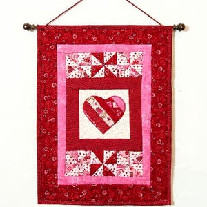 May include: A Valentine's Day wall hanging featuring a quilted design. The piece showcases a central heart motif, surrounded by pink and red borders. The red fabric has a swirl pattern. The hanging is approximately 12 inches wide and 18 inches long.