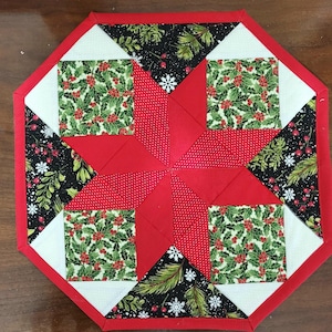 May include: A red, white, and green quilted table runner with a starburst design. The fabric features a holly pattern, a black and green floral pattern, and a red fabric with white polka dots.