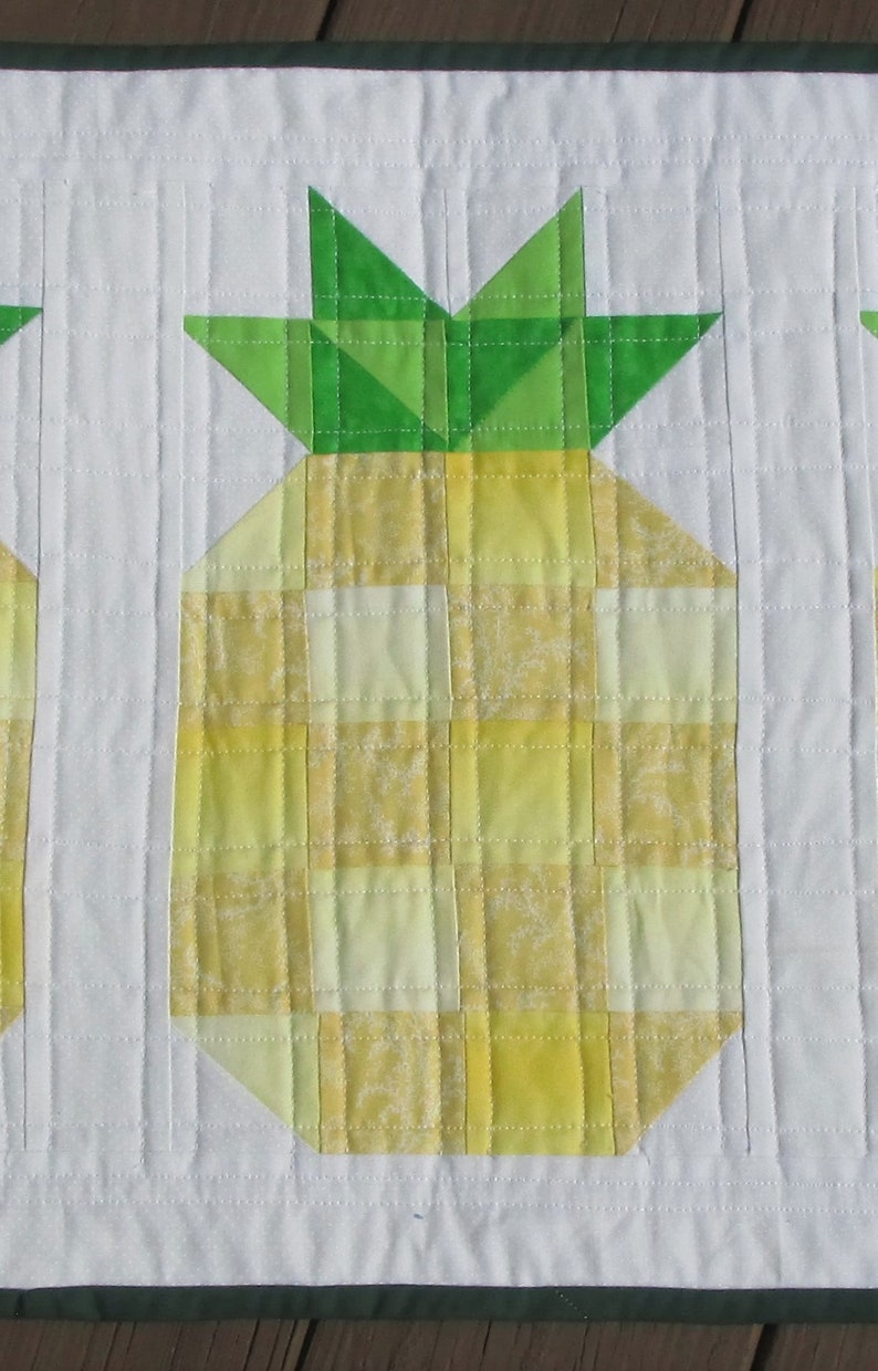 Puede incluir: Camino de mesa acolchado con un dise&ntilde;o de pi&ntilde;a repetido. Cada pi&ntilde;a est&aacute; hecha de tela de cuadros amarillos con hojas verdes, sobre un fondo blanco. Los bordes est&aacute;n acabados con un borde verde oscuro.