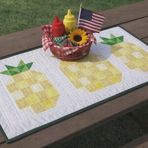 Puede incluir: Un camino de mesa acolchado blanco con tres dise&ntilde;os de pi&ntilde;as amarillas. Una cesta con ketchup, mostaza, un girasol y una peque&ntilde;a bandera americana se encuentra sobre el camino de mesa. El camino de mesa est&aacute; sobre una mesa de picnic de madera.