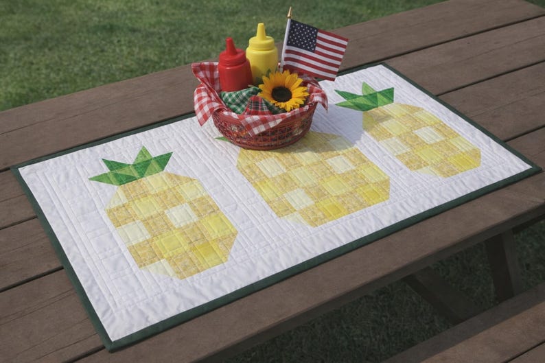 Puede incluir: Un camino de mesa acolchado blanco con tres dise&ntilde;os de pi&ntilde;as amarillas. Una cesta con ketchup, mostaza, un girasol y una peque&ntilde;a bandera americana se encuentra sobre el camino de mesa. El camino de mesa est&aacute; sobre una mesa de picnic de madera.