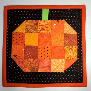 May include: A quilted Halloween-themed square featuring a pumpkin design. The pumpkin is made of orange fabric squares with various patterns. It has a green stem and is set against a black background with orange polka dots and a solid orange border.