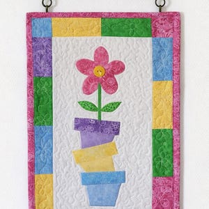 May include: A quilted wall hanging featuring a pink flower with a yellow button center, green leaves, and stacked flower pots in purple, yellow, and blue. The border is a patchwork of floral fabrics in pink, yellow, green, purple, and blue. Includes a decorative metal hanging bracket.