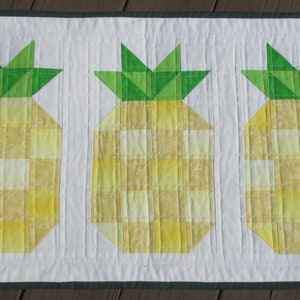 May include: A quilted table runner featuring three stylized pineapples. The pineapples have yellow and white checkered bodies and green tops. The runner is bordered with a dark green trim and white background.