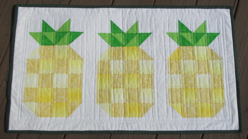 Puede incluir: Un camino de mesa acolchado con tres pi&ntilde;as estilizadas. Las pi&ntilde;as tienen cuerpos a cuadros amarillos y blancos y copas verdes. El camino de mesa tiene un borde verde oscuro y un fondo blanco.