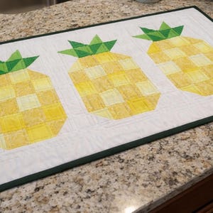 Puede incluir: Camino de mesa rectangular acolchado blanco con tres dise&ntilde;os de pi&ntilde;as amarillas y verdes. Las pi&ntilde;as tienen un patr&oacute;n de cuadros y copas verdes y frondosas. El camino de mesa est&aacute; bordeado con un ribete verde oscuro.