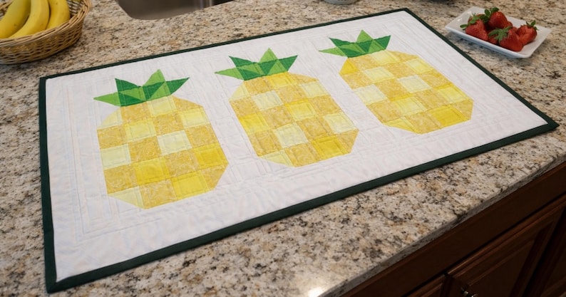 Puede incluir: Camino de mesa rectangular acolchado blanco con tres dise&ntilde;os de pi&ntilde;as amarillas y verdes. Las pi&ntilde;as tienen un patr&oacute;n de cuadros y copas verdes y frondosas. El camino de mesa est&aacute; bordeado con un ribete verde oscuro.